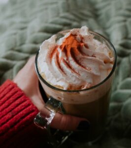 Une main tient une tasse de pumpkin spice latte surmonté de crème fouettée et de cannelle sous fond de plaid
