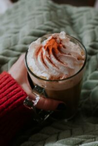 Une main tient une tasse de pumpkin spice latte surmonté de crème fouettée et de cannelle sous fond de plaid