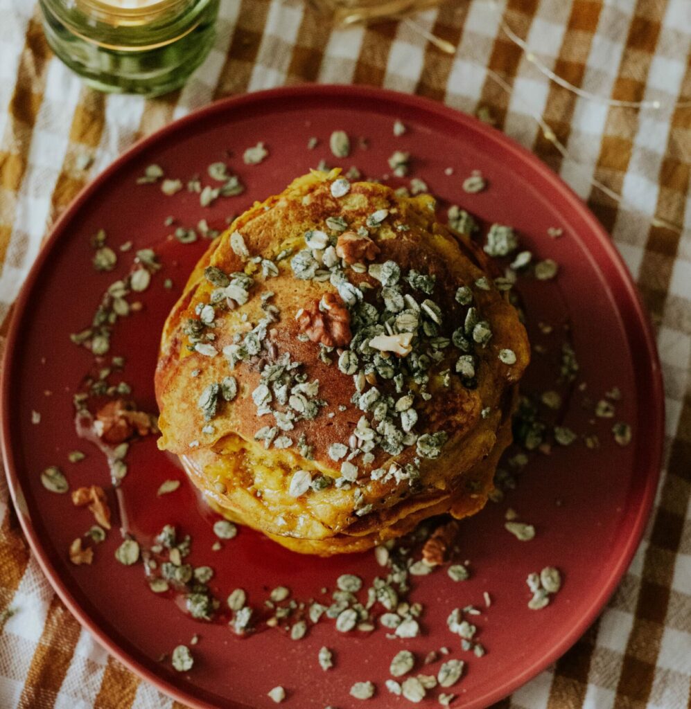 Pancakes à la citrouille vus de haut, servis sur une assiette terracotta, entourés de bougies et de muesli, sur un fond à carreaux orange aux tons automnaux.